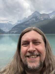 Smiling long haired man in front of a scenic lake